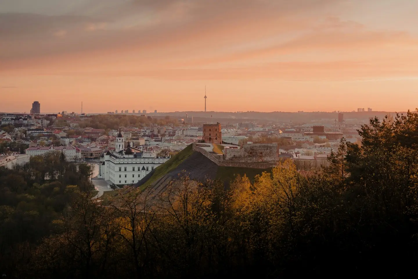 Rudeninė Vilniaus senamiesčio panorama saulėlydžio metu. Ant kalvos matyti Gedimino pilies bokštas ir pilies liekanos, o tolumoje driekiasi miesto pastatai po rausvu dangumi.