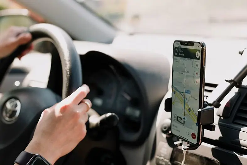 Automobilyje šalia vairo pritvirtintas išmanusis telefonas su atidaryta navigacijos programėle. Vairuotojo rankos laiko vairą kelionės metu.