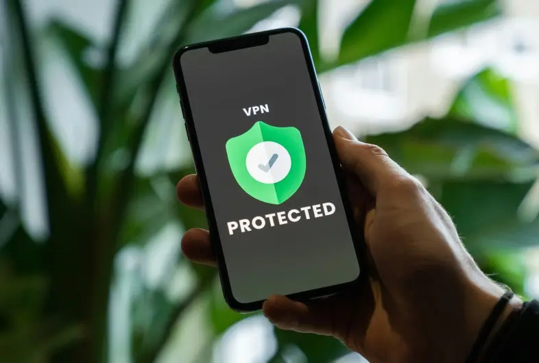 Rankoje laikomas išmanusis telefonas, kurio ekrane rodoma žalia VPN apsaugos piktograma ir užrašas „VPN Protected“. Fone matomi neryškūs žali augalai.