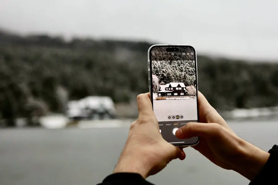 Mobilusis telefonas fotografuoja gyvenamąjį namą prie apsnigto miško, šalia pakrantės.