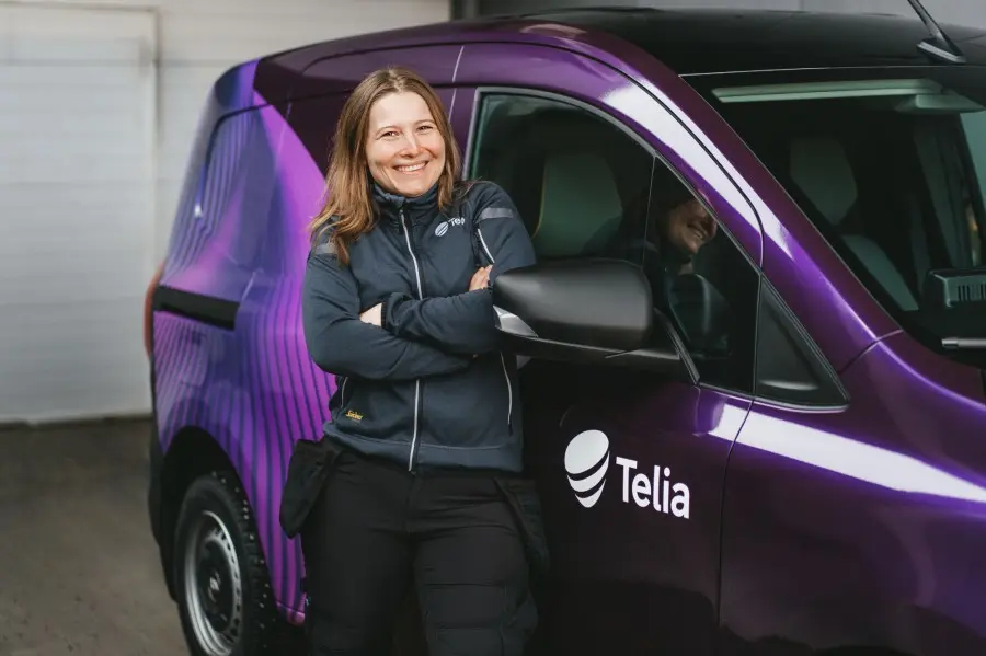 Telia inžinierė stovi prie Telia darbinio elektromobilio ir plačiai šypsosi