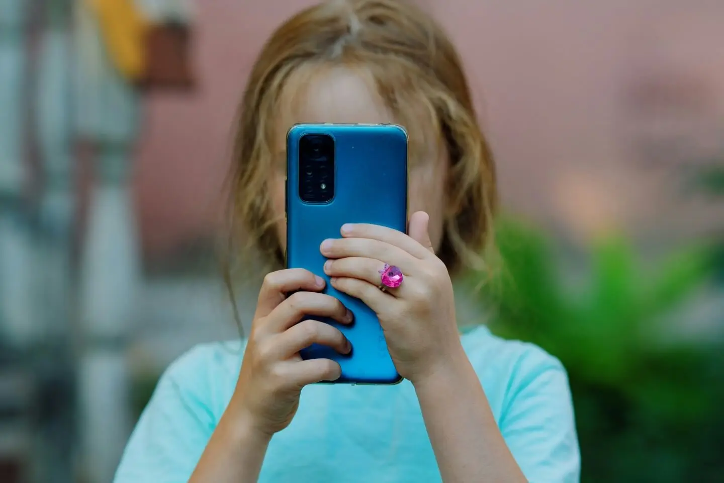 Mergaitė laiko mėlyną išmanųjį telefoną priešais veidą ir naudojasi juo lauke