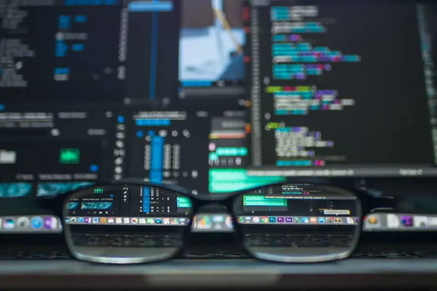Kompiuterio ekrane matomas programavimo kodas, atsispindintis ant akinių lęšių. Neryškus fonas pabrėžia programinės įrangos kūrimo ir technologijų aplinką.