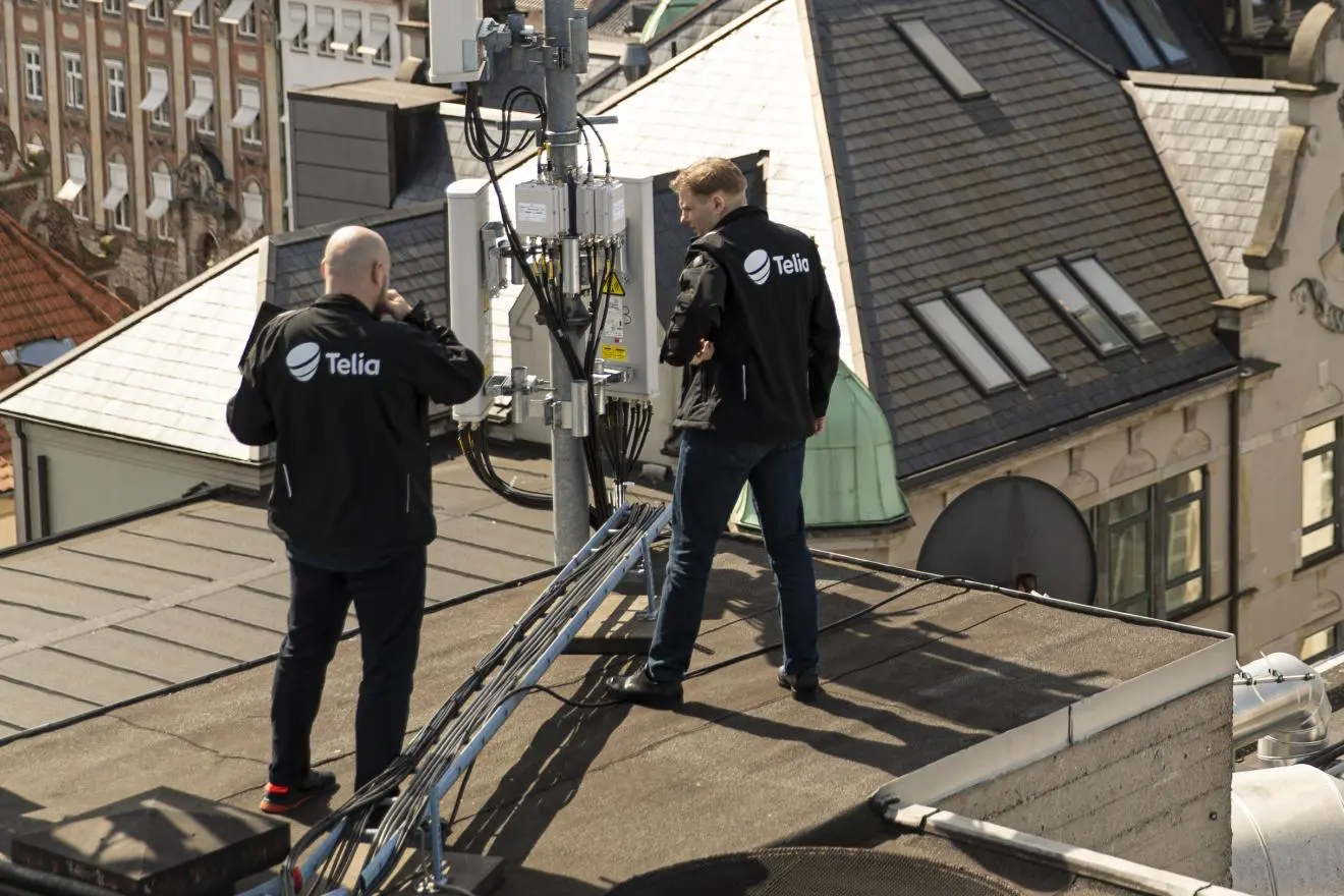 Du Telia darbuotojai ant pastato stogo apžiūri telekomunikacijų įrangą. Aplink matomi miesto pastatų stogai.