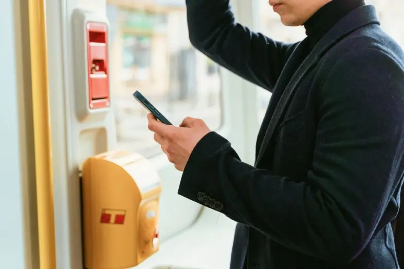 Asmuo viešajame transporte laiko išmanųjį telefoną prie bekontakčio bilietų ar mokėjimo skaitytuvo. Šalia matoma transporto priemonės vidaus įranga.