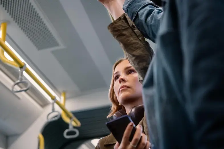 Mergina stovi viešojo transporto salone ir viena ranka laikosi už turėklo. Kitoje rankoje laiko išmanųjį telefoną. Matomos geltonos laikymosi rankenos.