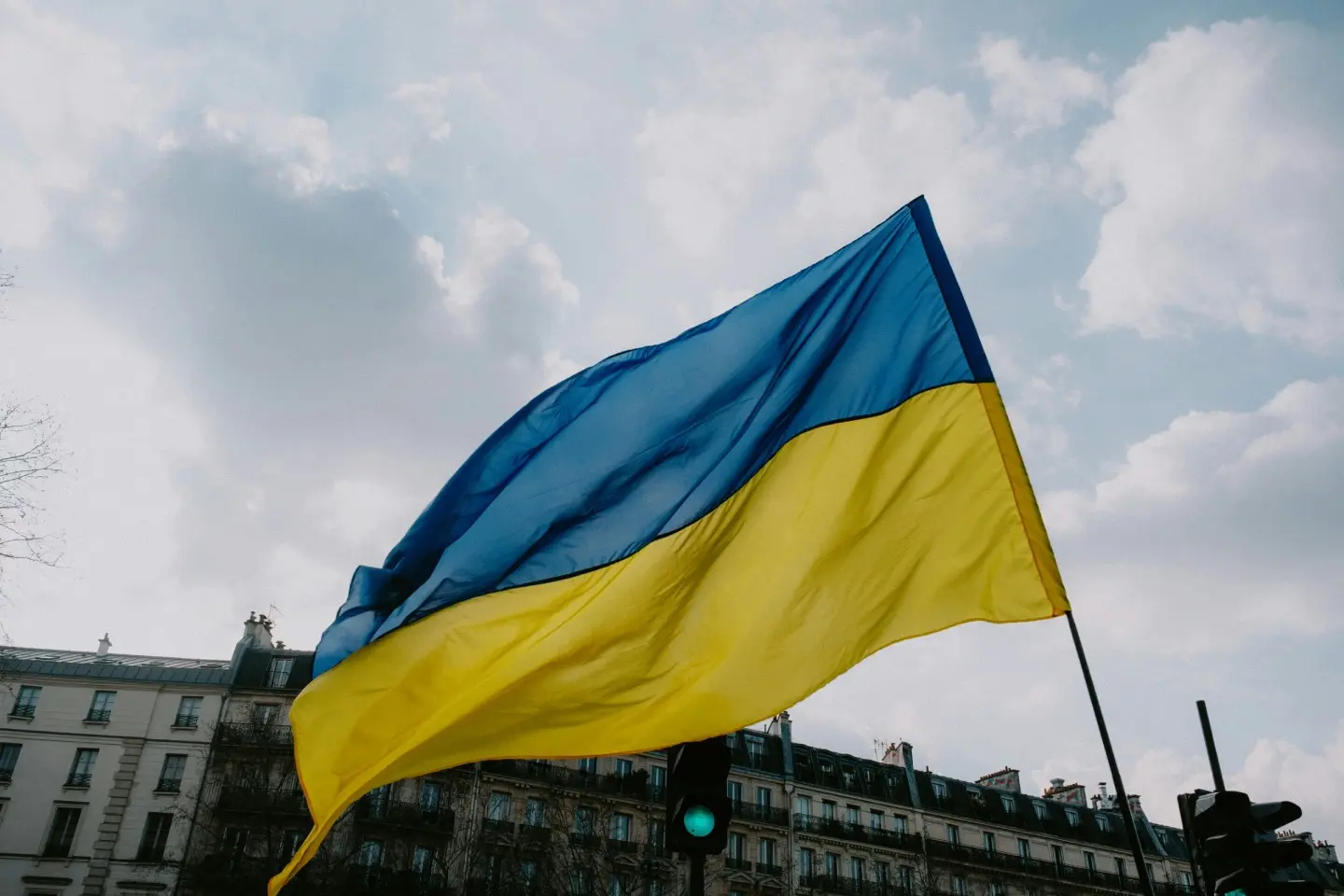 Aukštai į dangų iškelta Ukrainos vėliava. Už jos matosi debesuotas dangus ir senovinių pastatų viršus.