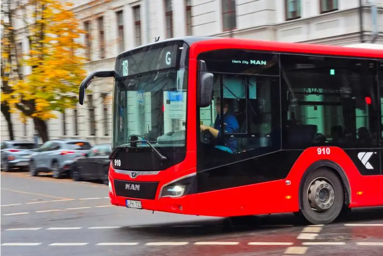 Raudonas miesto autobusas važiuoja miesto gatve pro sankryžą. Fone matomi pastatai ir kiti automobiliai.