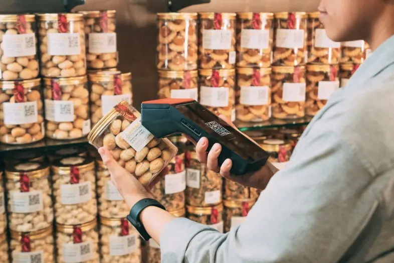 Asmuo parduotuvėje rankoje laiko riešutų stiklainį ir skeneriu nuskaito QR kodą ant etiketės. Lentynose už nugaros išrikiuoti panašūs produktai.