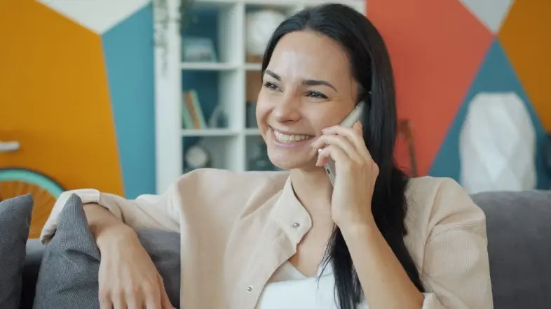 Moteris sėdi ant sofos ir kalba mobiliuoju telefonu. Fone matoma spalvinga geometrinių formų siena ir lentyna su daiktais.