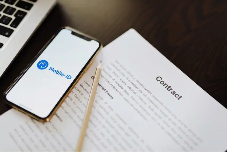 Ant stalo padėtas išmanusis telefonas su „Mobile-ID“ programėlės ekranu. Šalia matomas rašiklis ir atverstas sutarties dokumentas su antrašte „Contract“.