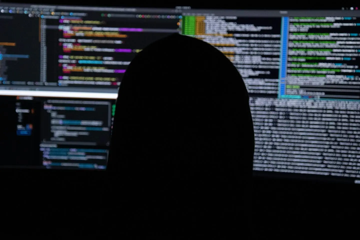 Asmuo stebi kelis kompiuterių ekranus, kuriuose rodomas programinis kodas, sistemos duomenys ir techninė informacija. Vaizdas siejamas su kibernetiniais nusikaltėliais
