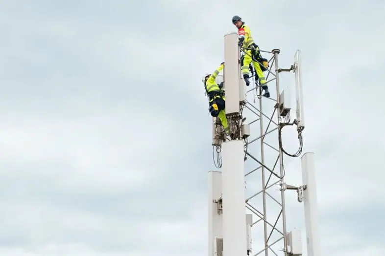 Du techninės priežiūros darbuotojai su apsaugine apranga dirba mobiliojo ryšio bokšto viršuje. Matomos antenos ir debesuotas dangus fone.