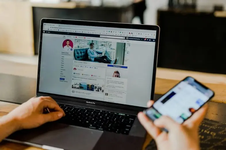 Nešiojamas kompiuteris ant stalo su atvertu Facebook profiliu, kitoje rankoje žmogus laiko telefoną.