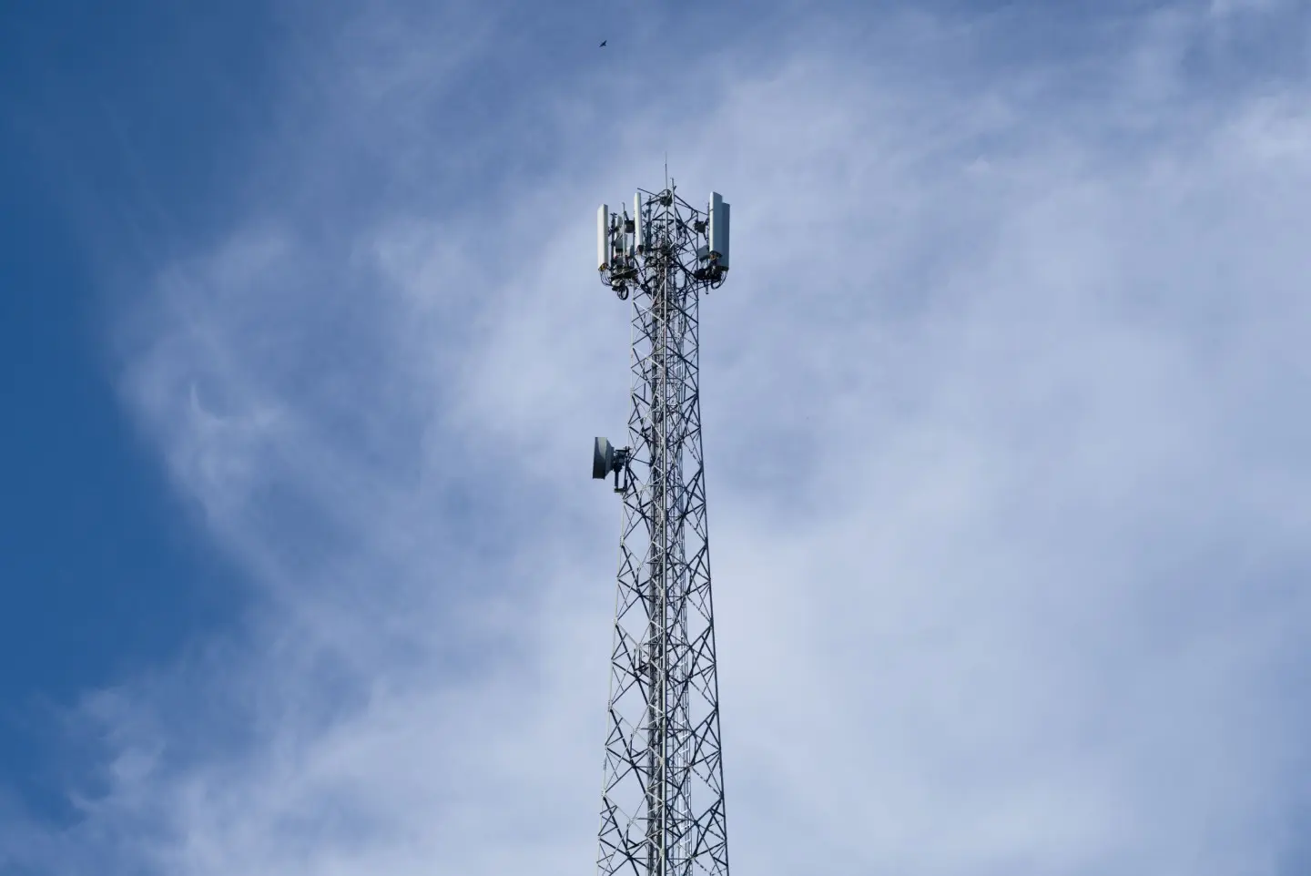 Mobiliojo ryšio bokšto viršūnė aukštai žydrame danguje su keliais debesėliais.