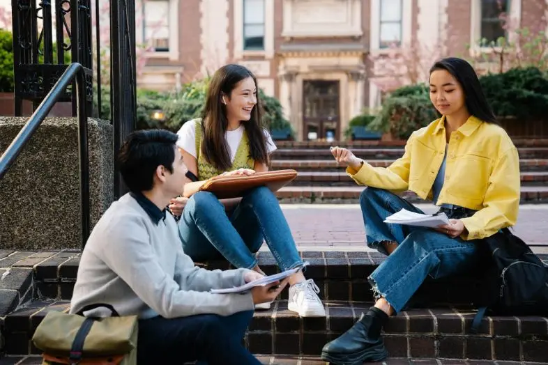 Trys besimokantys asmenys susėdę ant laiptų universiteto kieme. Jie kalbasi ir peržiūri užrašus, ruošdamiesi ar aptardami studijų užduotis.