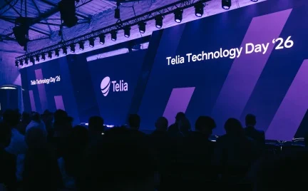 „Telia Technology Day ’26“ renginio scena (fotografė Armida Grosaitė) su dideliais LED ekranais ir „Telia“ logotipu, apšviesta violetine šviesa. Priekyje matoma auditorija, stebinti pristatymą technologijų konferencijoje.