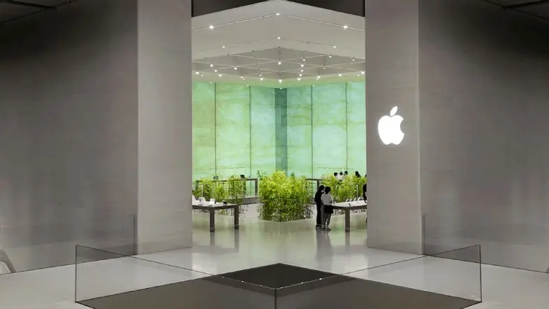 Modernios „Apple“ parduotuvės interjeras su dideliu šviečiančiu „Apple“ logotipu. Erdvioje salėje matomi produktų stalai, augalai ir šviesiai žalias fonas.