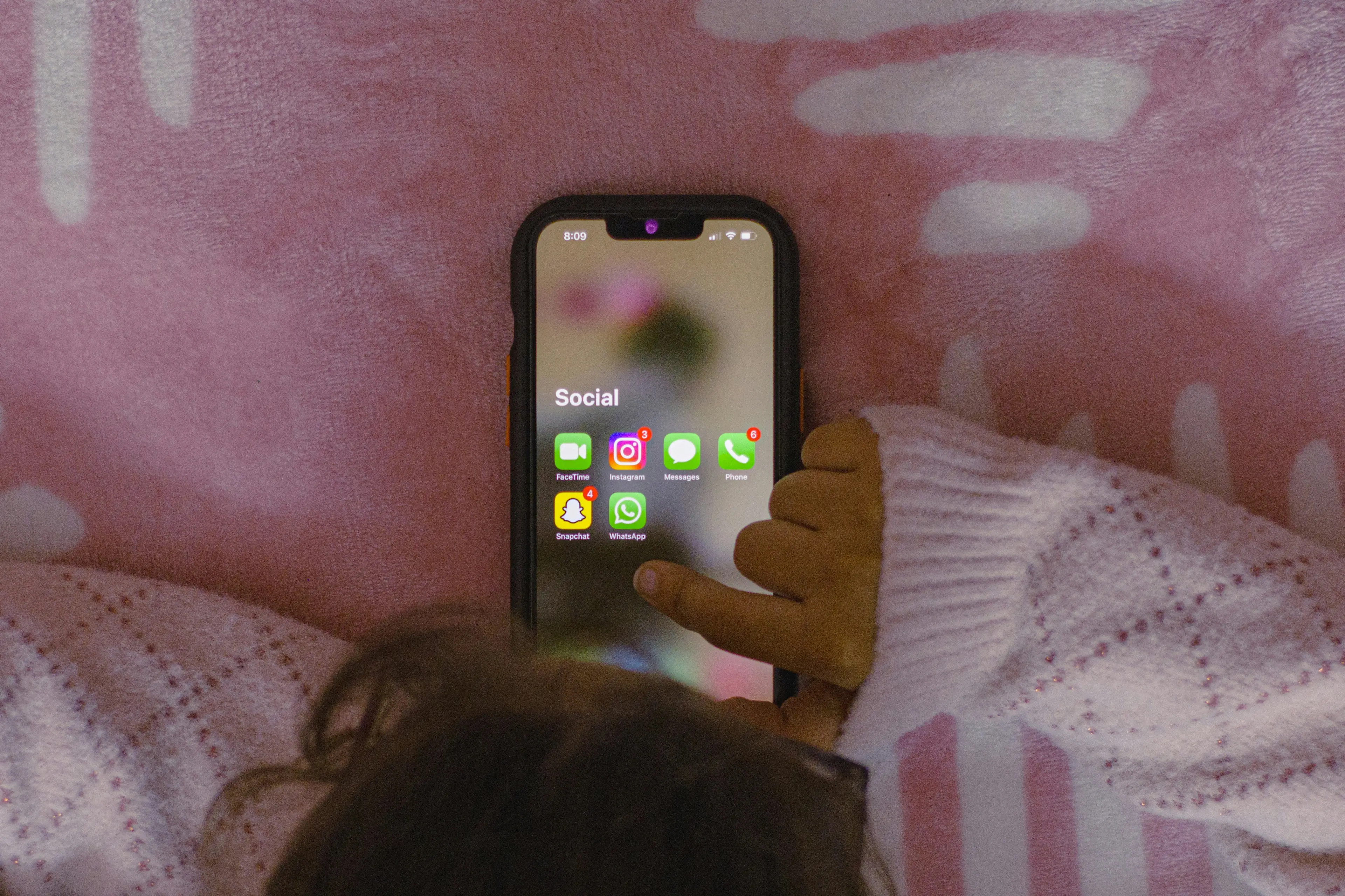 Iš viršaus matosi telefonas ant lovos su socialinėm programėlėm ir vaiko ranka, kuri ruošiasi spausti programėlę.
