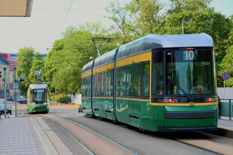 Žalias miesto tramvajus važiuoja bėgiais medžių apsuptoje gatvėje. Tolumoje matomas dar vienas tramvajus, šaligatvis ir miesto pastatai.
