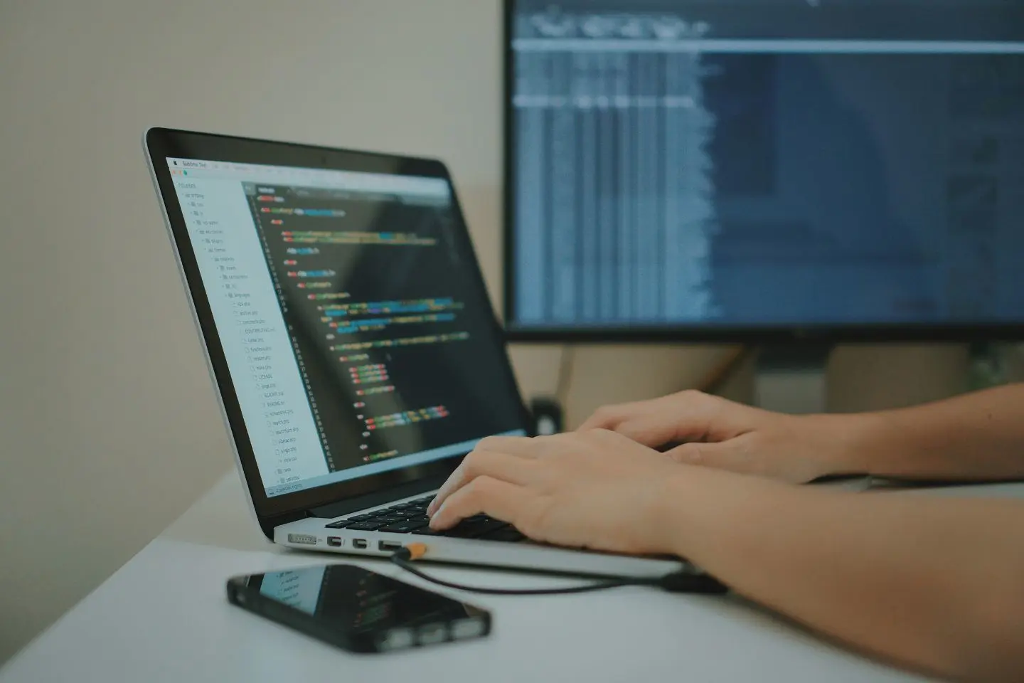 Asmuo dirba prie nešiojamojo kompiuterio. Ekrane matomas programavimo kodas, o fone stovi antras monitorius su atverta programine įranga.