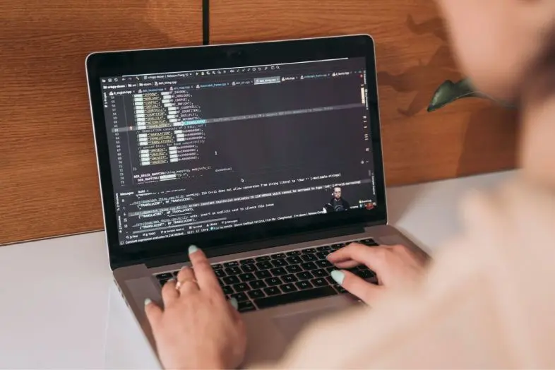 Asmuo sėdi prie stalo ir rašo nešiojamame kompiuteryje, kurio ekrane yra programavimo kodas.