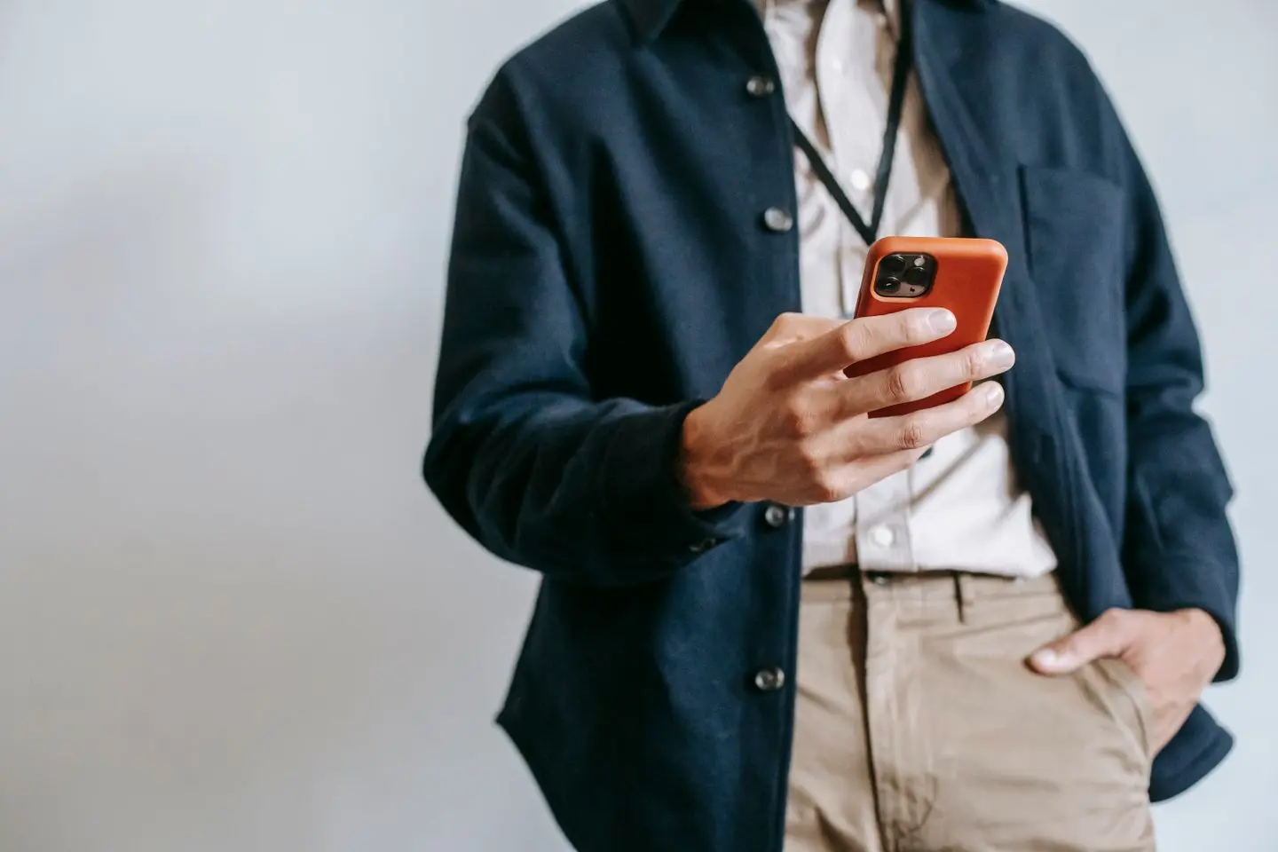 Asmuo stovi ir žiūri į išmanųjį telefoną oranžiniame dėkle. Matoma tamsi striukė, šviesūs marškiniai ir ranka kišenėje.