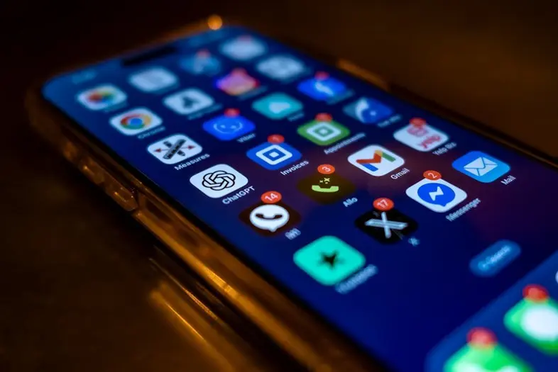 Iš arti nufotografuotas išmaniojo telefono ekranas su daugybe programėlių piktogramų. Tarp jų matomos „ChatGPT“, „Gmail“, „Messenger“ ir kitos programos.