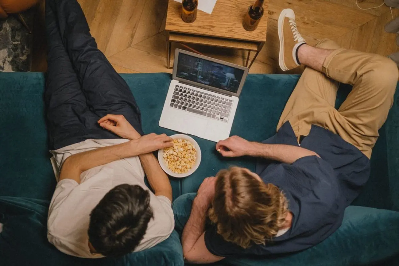 Vaizdas iš viršaus: du žmonės sėdi ant sofos ir žiūri filmą nešiojamajame kompiuteryje. Tarp jų dubuo spragėsių, šalia staliukas su gėrimų buteliais.