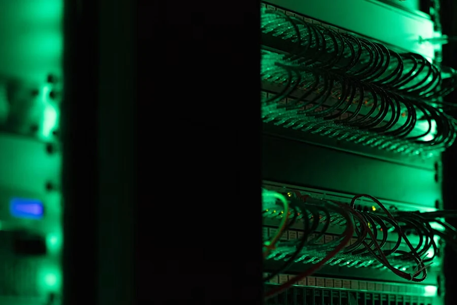 Serverių spinta su tinklo įranga ir tankiai sujungtais kabeliais. Žalias apšvietimas pabrėžia duomenų centro technologinę aplinką.