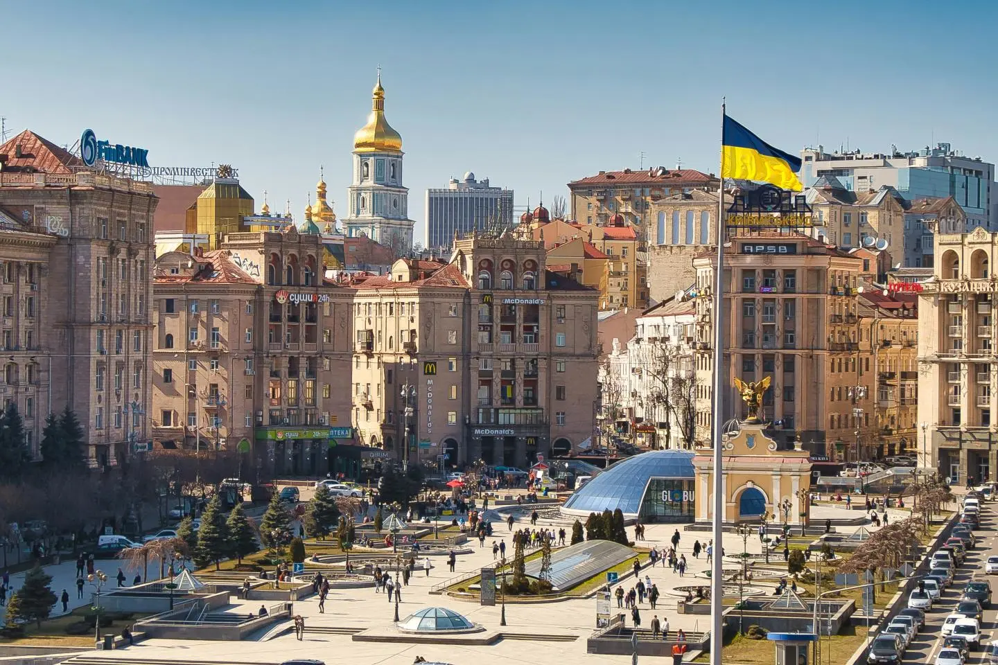 Kyjivo centrinės aikštės panorama saulėtą dieną. Virš miesto pastatų plevėsuoja didelė Ukrainos vėliava, fone matyti bažnyčių bokštai ir miesto architektūra.