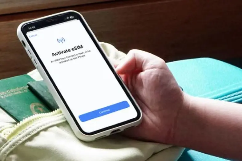 Asmuo rankoje laiko išmanųjį telefoną su atidarytu eSIM aktyvavimo langu. Telefonas padėtas ant kelioninio krepšio šalia paso.