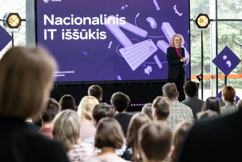 Renginio metu prieš susirinkusią auditoriją vyksta pristatymas. Scenoje šalia mikrofono stovi pranešėjas, o ekrane rodomas užrašas „Telia Nacionalinis IT iššūkis“.