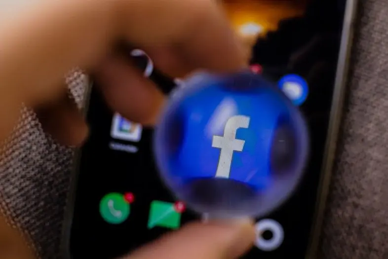 Per padidinamąjį stiklą matomas „Facebook“ logotipas išmaniojo telefono ekrane. Fone neryškiai matomos kitos programėlių piktogramos.