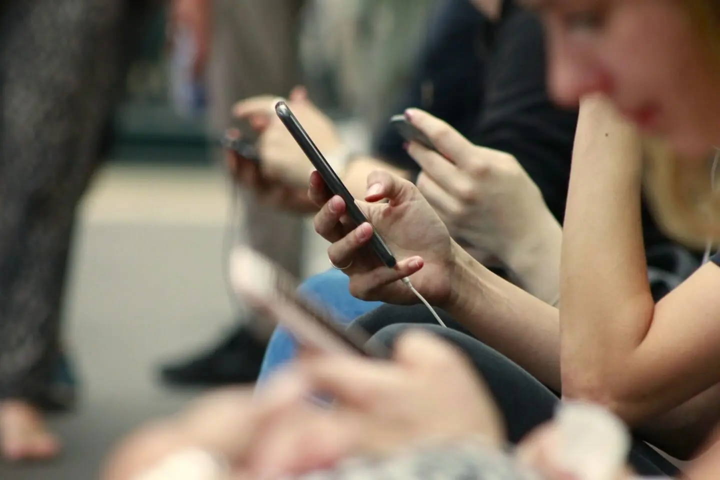 Žmonės sėdi viešoje vietoje ir žiūri į savo telefonus, kiekvienas susikoncentravęs į ekraną, fone matoma neryški aplinka.