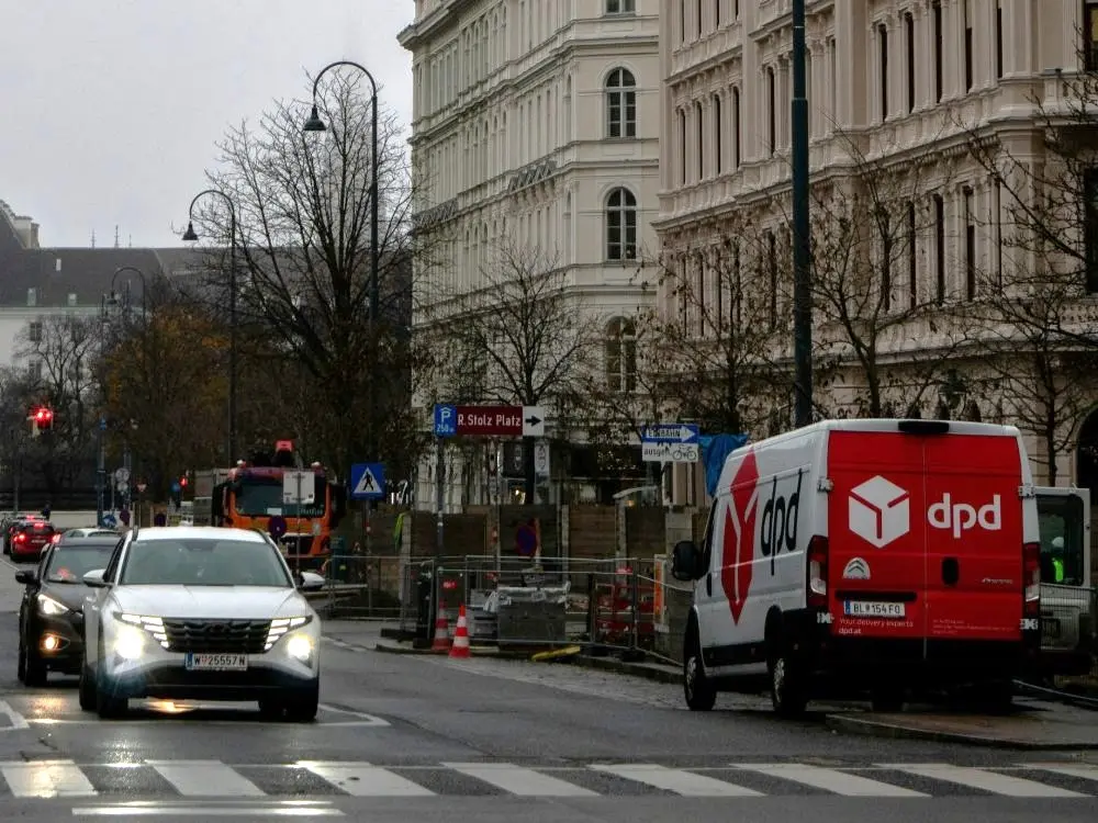 Šlapia miesto gatvė tarp istorinių daugiaaukščių pastatų. Dešinėje stovi raudonai baltas DPD kurjerių furgonas, o kairėje važiuoja automobiliai su įjungtais žibintais.