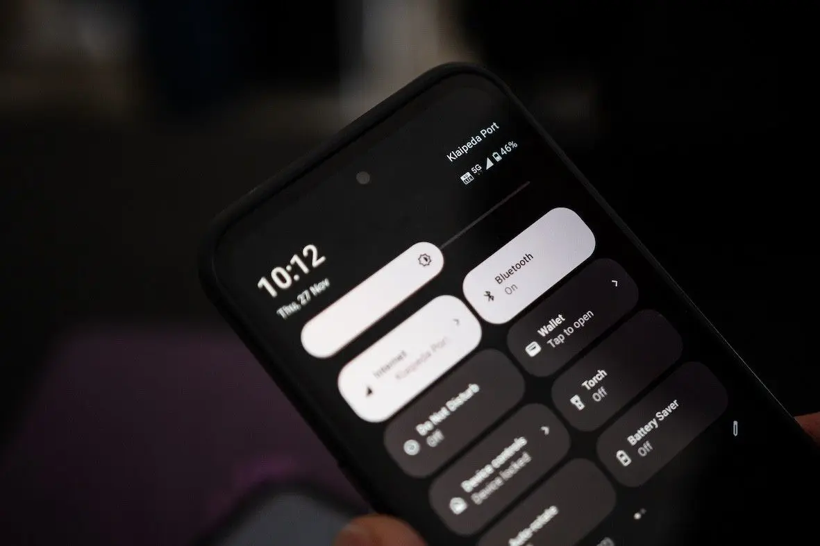 Iš arti nufotografuotas išmanusis telefonas tamsiame fone. Ekrane atidarytas greitųjų nustatymų meniu su „Bluetooth“, žibintuvėlio ir baterijos taupymo parinktimis.