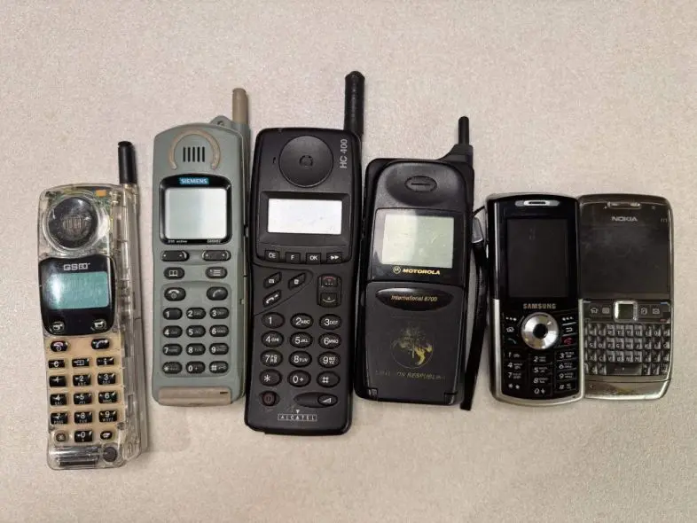 Seni namų telefonai ir mobilieji telefonai guli ant grindų, išdėlioti eile.