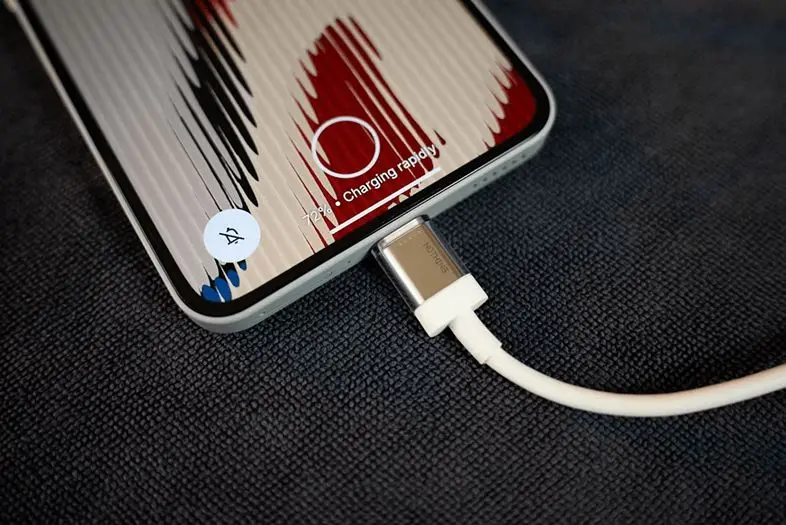 Išmanusis telefonas guli ant tamsaus audinio paviršiaus ir yra prijungtas prie balto įkrovimo laido. Ekrane matomas greitojo įkrovimo indikatorius ir 12 % baterijos lygis.