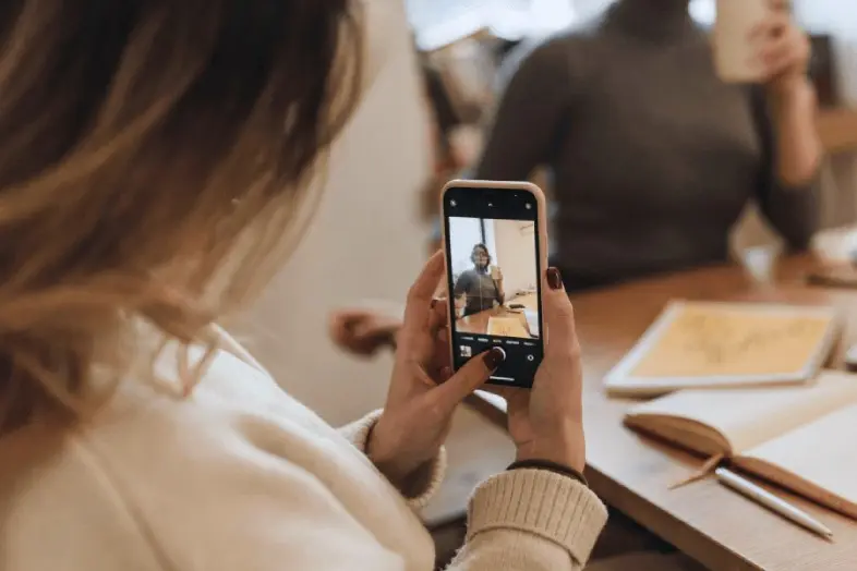 Viena moteris laiko išmanųjį telefoną ir fotografuoja kitą moterį, sėdinčią prie stalo. Ant stalo matomi dokumentai, planšetė ir rašiklis.