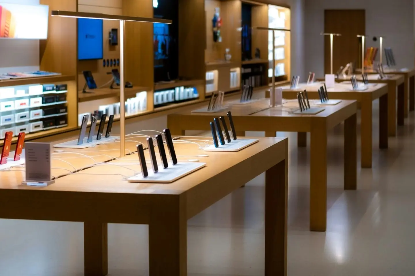 Elektronikos parduotuvės salonas su keliais mediniais stalais, ant kurių išdėstyti demonstraciniai išmanieji telefonai. Šviesi ir tvarkinga ekspozicijos erdvė.