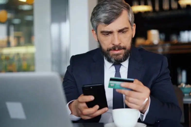 Asmuo prie nešiojamojo kompiuterio vienoje rankoje laiko telefoną, kitoje banko kortelę. Vaizduojamas apsipirkimas ar mokėjimo operacijos atlikimas internetu.