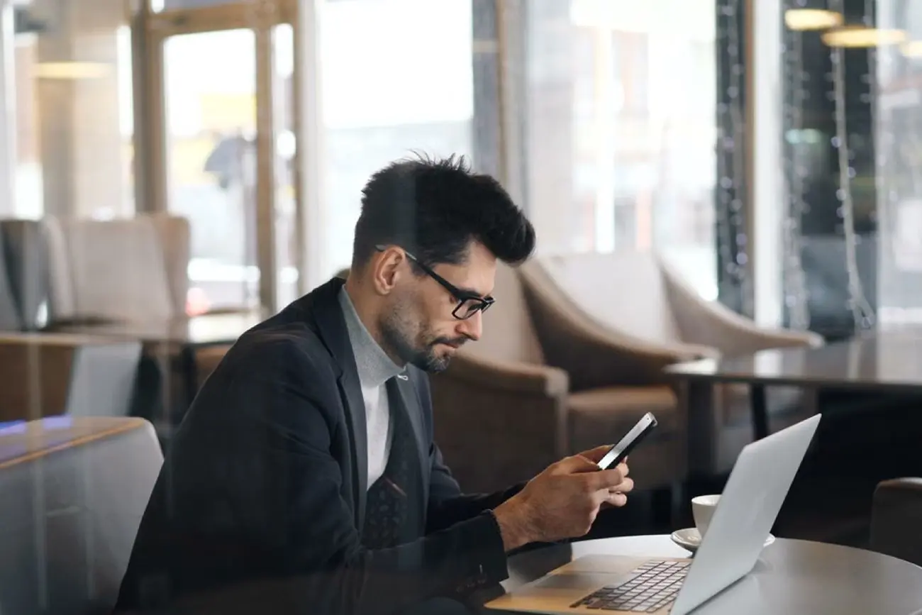 Vyras sėdi prie stalo kavinėje ar poilsio erdvėje, rankoje laiko išmanųjį telefoną. Ant stalo priešais atidarytas nešiojamasis kompiuteris ir puodelis.