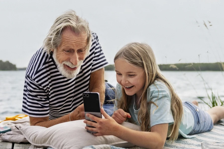 Mergaitė ir senelis guli ant medinio liepto prie ežero ir kartu žiūri į išmanųjį telefoną