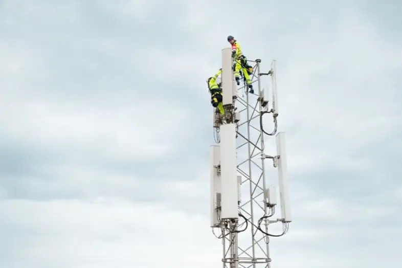Du techninės priežiūros darbuotojai, vilkintys ryškias apsaugines striukes, dirba aukštai ant mobiliojo ryšio bokšto. Fone matyti apsiniaukęs dangus.