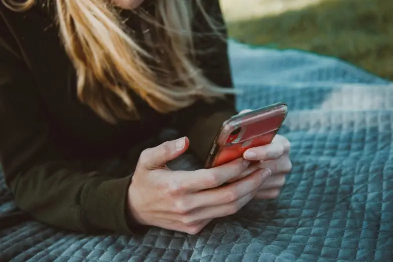 Asmuo, sėdėdamas ant užkloto, abiem rankomis laiko išmanųjį telefoną. Matoma tik dalis kūno ir telefono.