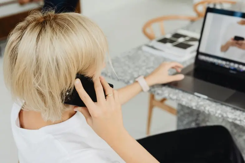 Asmuo sėdi prie stalo, kalba mobiliuoju telefonu ir naudojasi nešiojamuoju kompiuteriu.