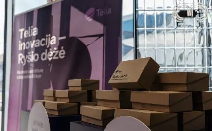 Ant stalo sukrautos dėžutės prie stendo su užrašu „Telia inovacija – Ryšio dėžė“. Vaizde matoma kampanijos ar renginio ekspozicija.