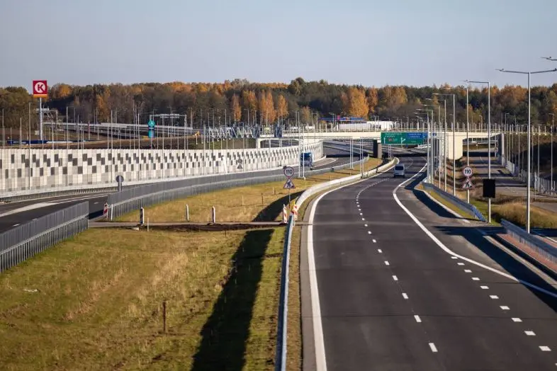 Platus naujai įrengtas greitkelis su keliomis eismo juostomis, viaduku ir kelio ženklais. Fone matyti rudeninių spalvų miškas ir pavienės transporto priemonės.