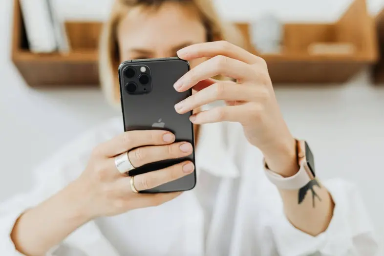 Moteris su baltais marškiniais laiko išmanųjį telefoną abiem rankomis ir žiūri į ekraną. Ant jos rankos yra išmanusis laikrodis. Fonas neryškus.
